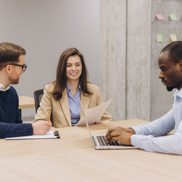 A diverse group of professionals seated at a table, one writes on a clipboard, another reviews a sheet of paper, the third types on a laptop